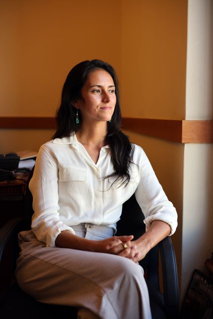 A person in a white shirt sits in a chair by the window, gazing thoughtfully to the side.