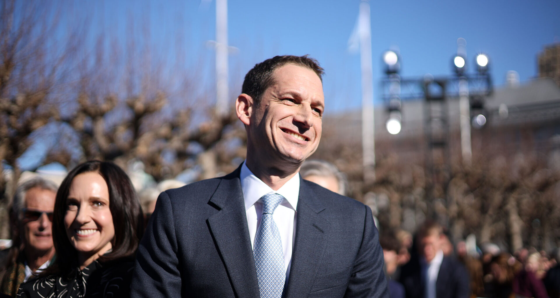 A man in a suit smiles while standing outdoors among a group of people under a clear blue sky.
