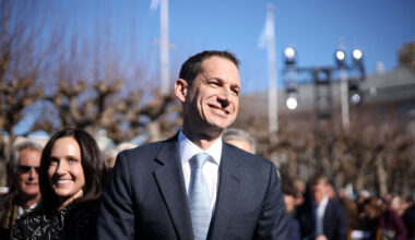 A man in a suit smiles while standing outdoors among a group of people under a clear blue sky.