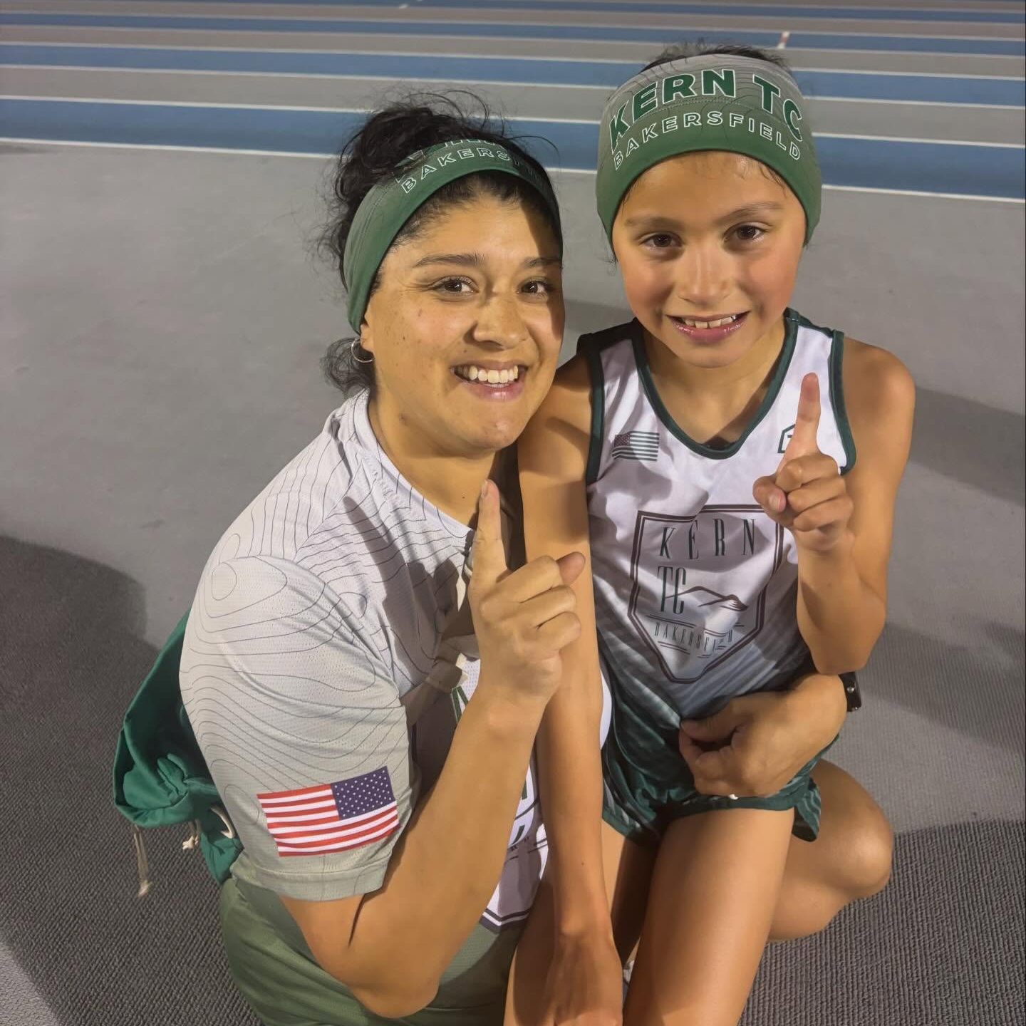 Emelie Chavez, the 7-year-old world record holder in the 3000M, and an adult pose for a picture on a track, both holding up one finger to signify their accomplishment.