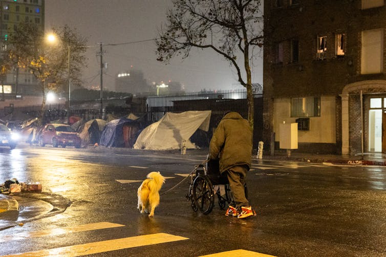 A homeless man walks a dog toward a group of tents lining a sidewalk.