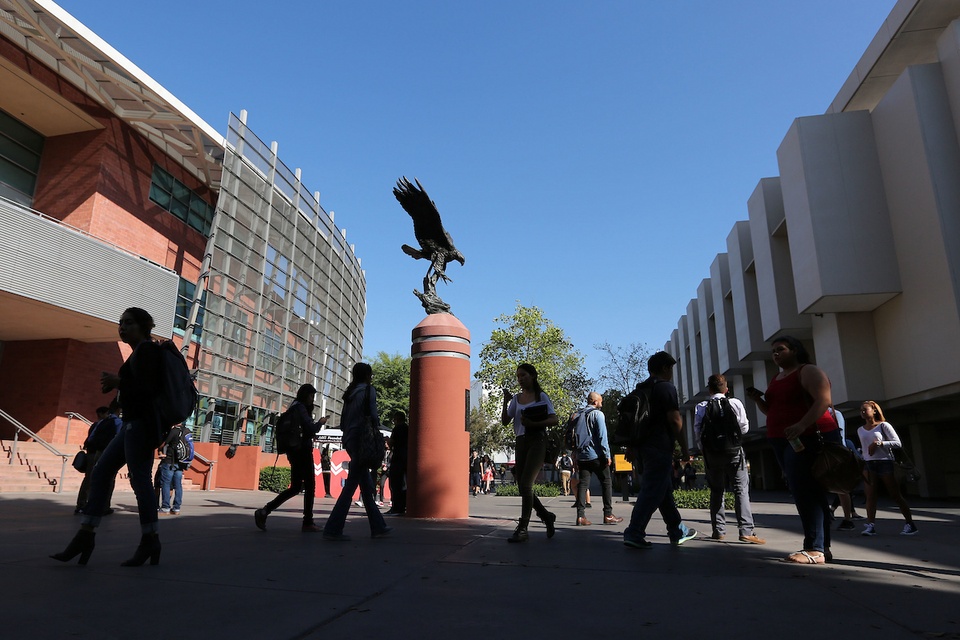 Students walk across campus beneath the Golden Eagle statue at Cal State LA, with modern buildings and a clear blue sky overhead.