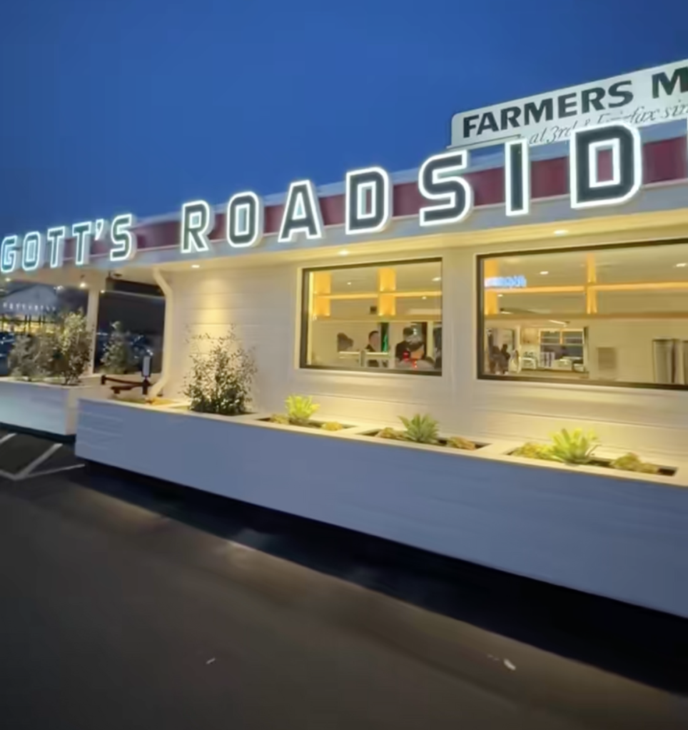 Exterior of Gott's Roadside restaurant with "Gott's Roadside" and "Farmers Market" signs illuminated at dusk.