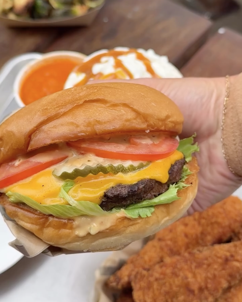 Close-up of a hand holding a cheeseburger with lettuce, tomato, pickles, and sauce.