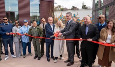 Group of officials and community members participate in a ribbon-cutting ceremony outside a modern building, with a red ribbon stretched across the front.
