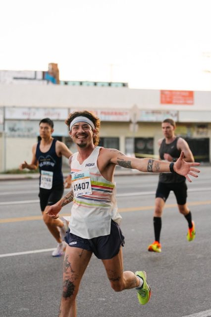 A smiling runner with arms outstretched leads two others in a race on a city street, wearing a race bib and athletic gear.