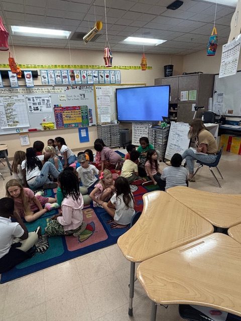 Youngsters sit in a classroom with art on the walls and a television screen behind them.