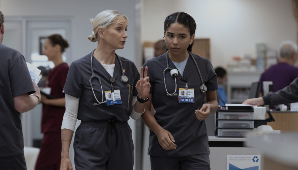 Two nurses, played by Katherine LaNasa and Laetitia Hollard, walk through a busy hospital corridor in scrubs. One gestures while speaking, capturing a collaborative moment on the set of The Pitt.
