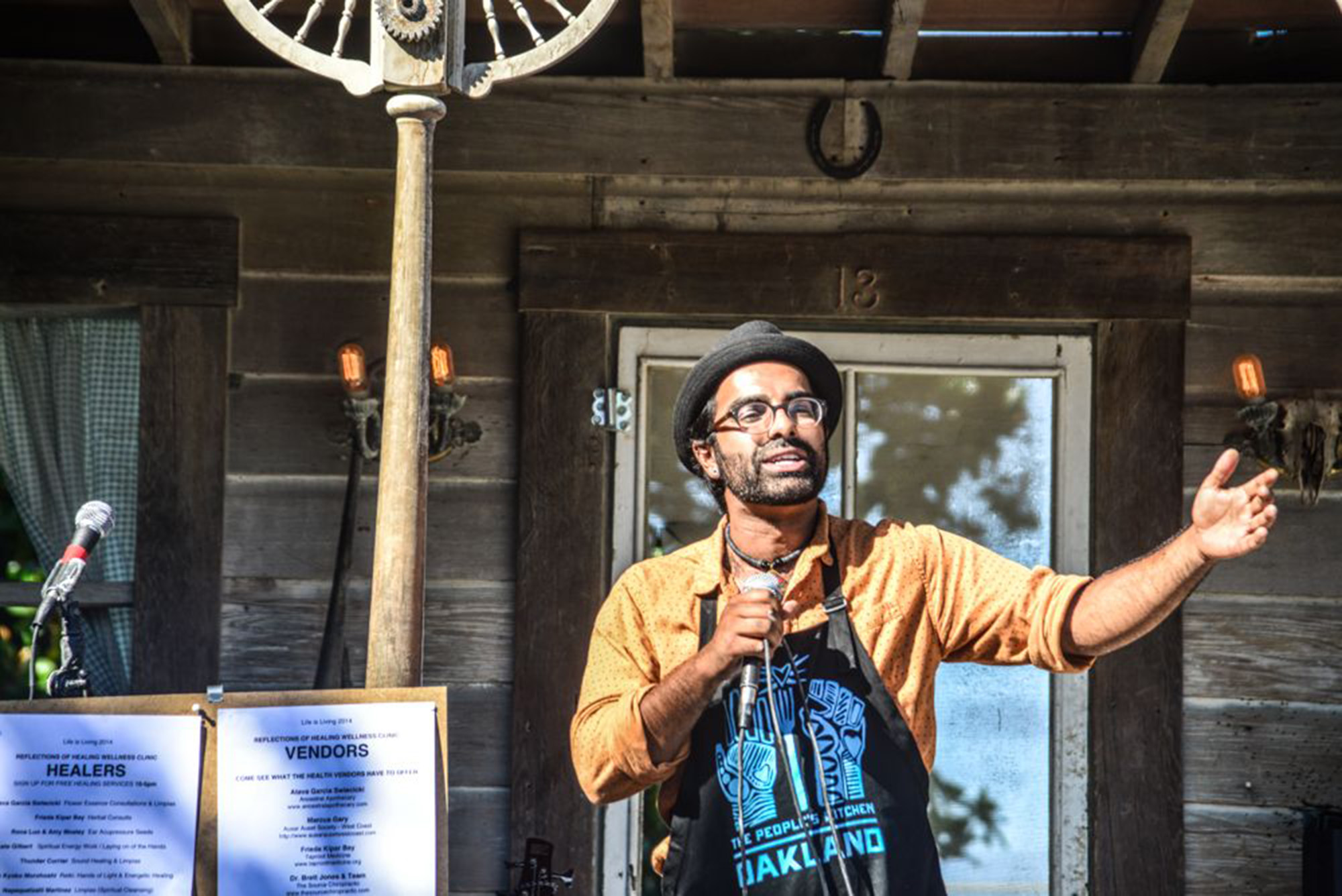 A South Asian man holds a microphone as he addresses a gathering. His apron reads, "The People's Kitchen, OAKLAND."