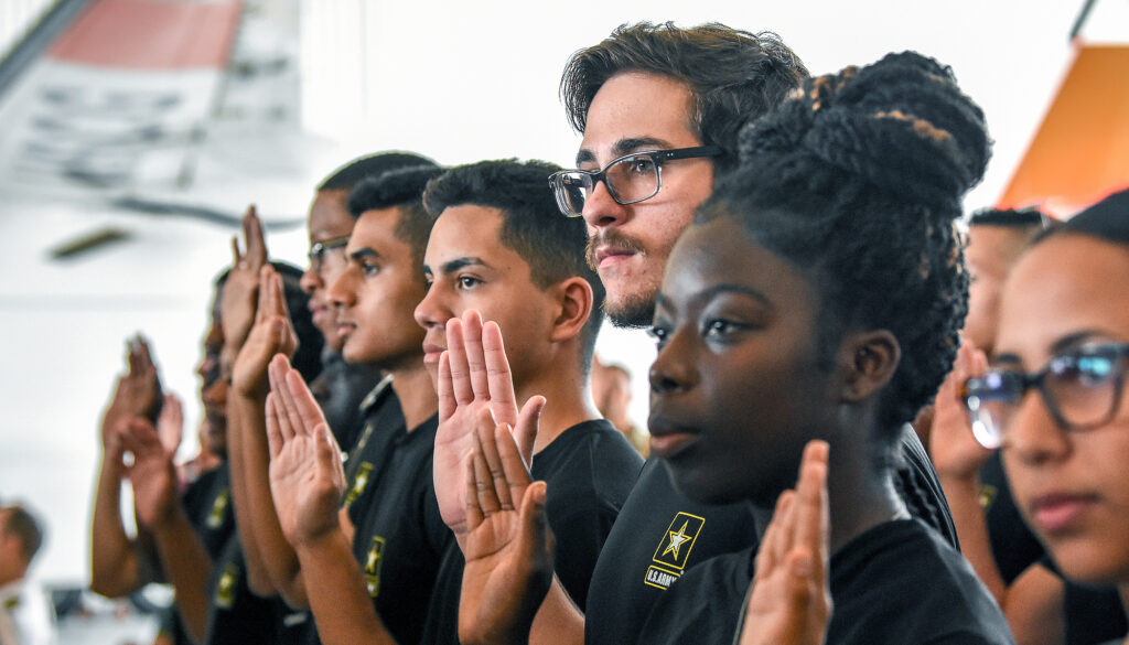 A line of young U.S. Army recruits, men and women of different races and ethnic backgrounds, stands in a line with right hands raised.