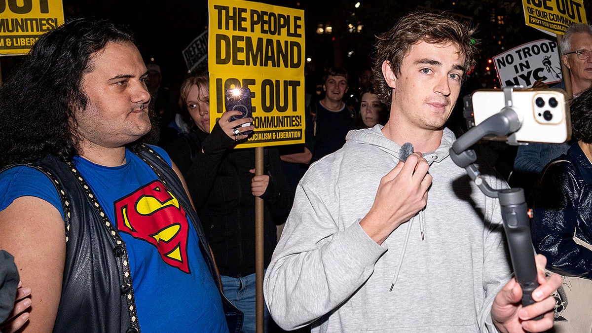 Nick Shirley holds a camera while filming protesters gathered during a demonstration on a city street.