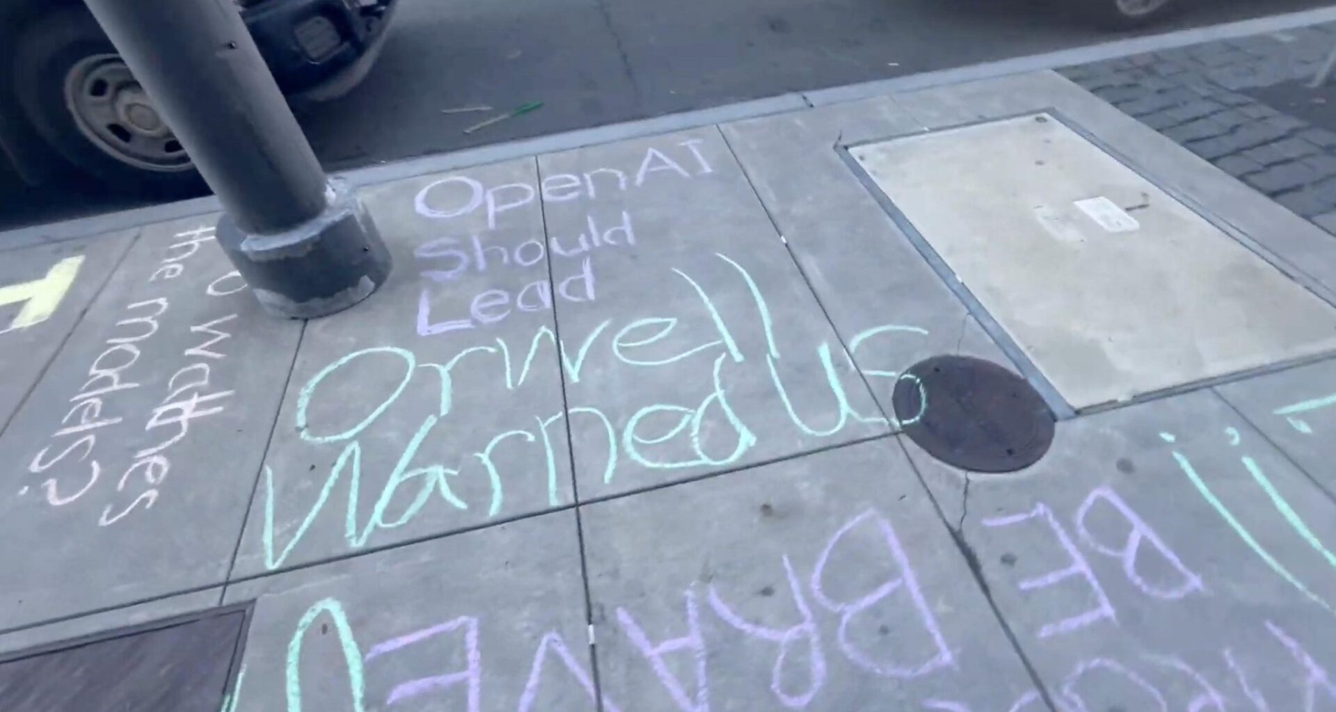 Chalk writing on a sidewalk reads, "OpenAI Should Lead Orwell Warned Us," with additional partially visible words and passing cars nearby.