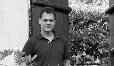 Black-and-white portrait of a man standing beside a wooden gate and stucco wall, surrounded by foliage.