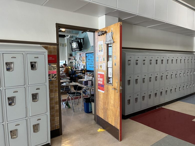 A teacher sits inside an empty classroom, viewed from the hallway of a school.