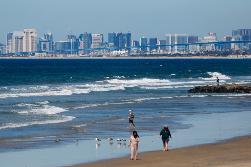 People walking and a man fishing on Imperial Beach with downtown San Diego and a bridge in the background.