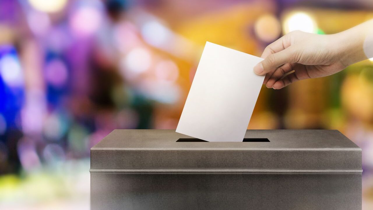 Colorful image of a hand, a white sheet of paper, and a box symbolizing the act of voting