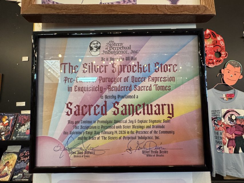 A framed certificate proclaims The Silver Sprocket Store as a Sacred Sanctuary, awarded by The Sisters of Perpetual Indulgence, displayed on a wall among t-shirts and other items.
