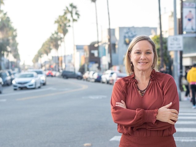 A photo of Traci Park in a maroon top smiling in front of a palm tree lined street with cars.