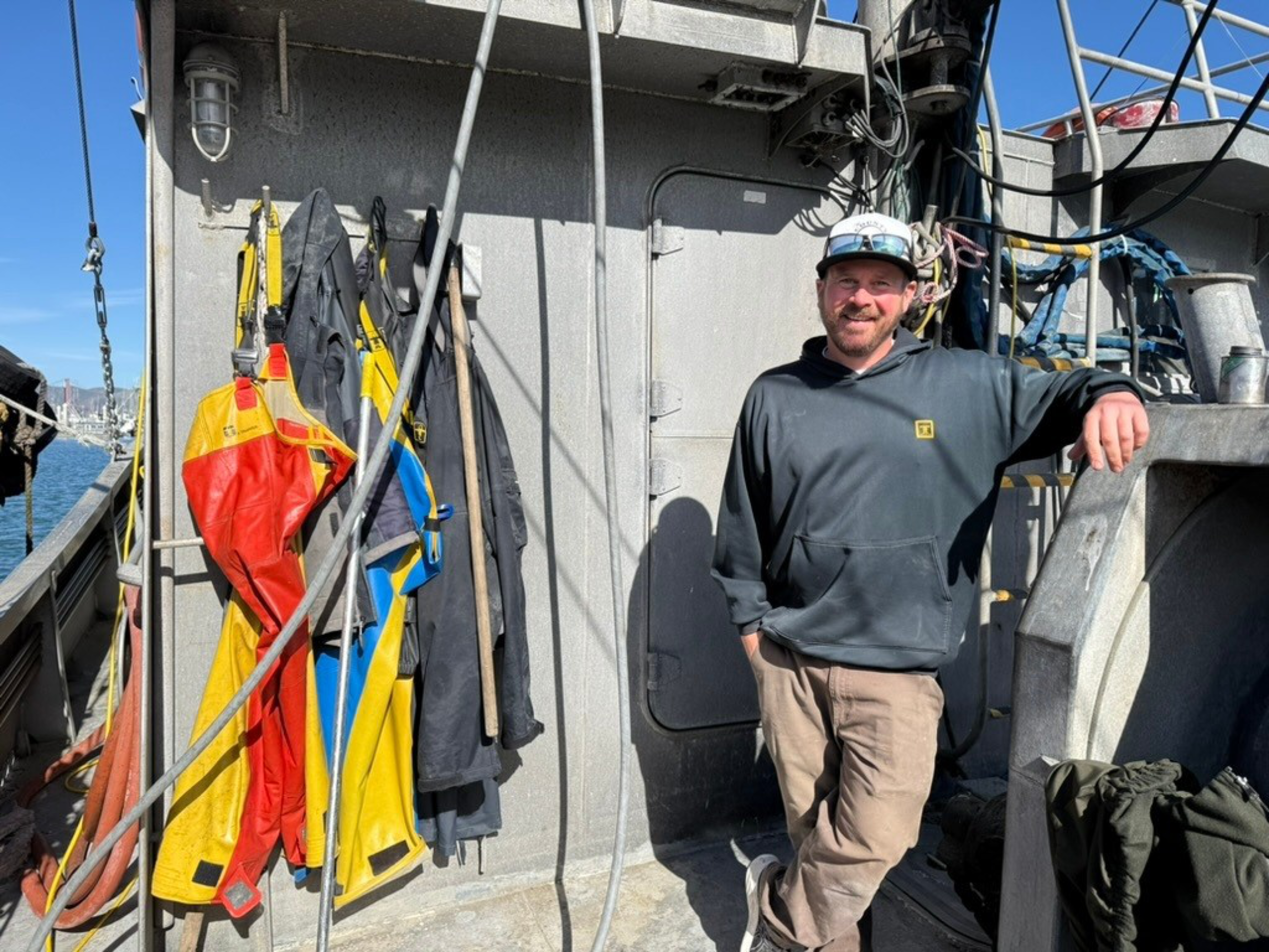 A man in a black hoodie and cap leans against a gray wall on a boat, with colorful rain jackets and fishing gear hanging nearby.