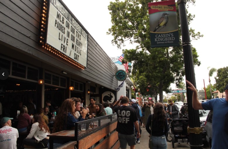 A small marquee for the Rabbit Hole bar advertises baseball and live music. The sign hangs over a patio filled with people and as passersby walk on the sidewalk near a post with a banner for Adams Avenue.