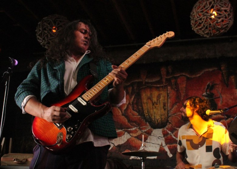 A long-haired man playing guitar stands on stage with a drummer in the background, half in the spotlight.