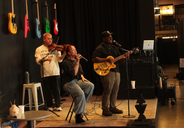 Three people perform on a small stage against a wall decored by five hanging guitars. One, with a microphone is seated on a stool. The man in back plays a violin and another man, in front, plays guitar while singing into a standing mic.