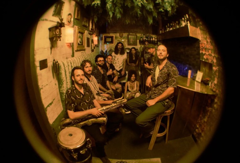 A man sits on a tall stool with four others lounging across from him, one of them holding a saxophone. One woman crouches behind him along with two other men and another woman. Greenery hangs above them all.