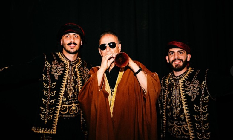 Three men stand, the one in the center in robes while playing an instrument, the other two in black jackets adorned with intricate gold embroidery.