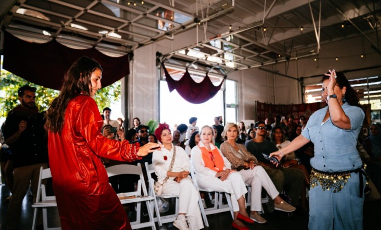 An audience sits on folding chairs in a large space watching as two people dance in the front of the room.