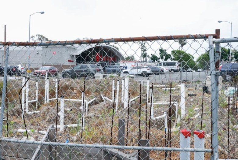 A Target store can be seen through a chain link fence around a foundation with poles sticking up from it that is overrun by weeds.