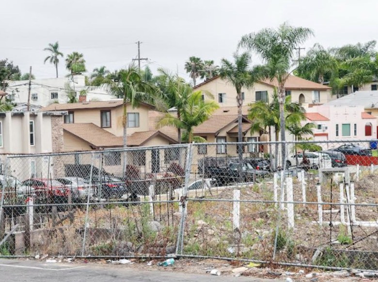 A line of homes on a street behind a parking lot can be seen through a leaning chain-link fence around a foundation that is overgrown with weeds.