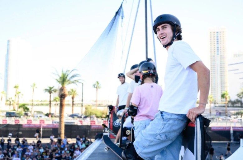 A man in a helmet leans against a railing with a sail on display behind him and a line of children to his side.
