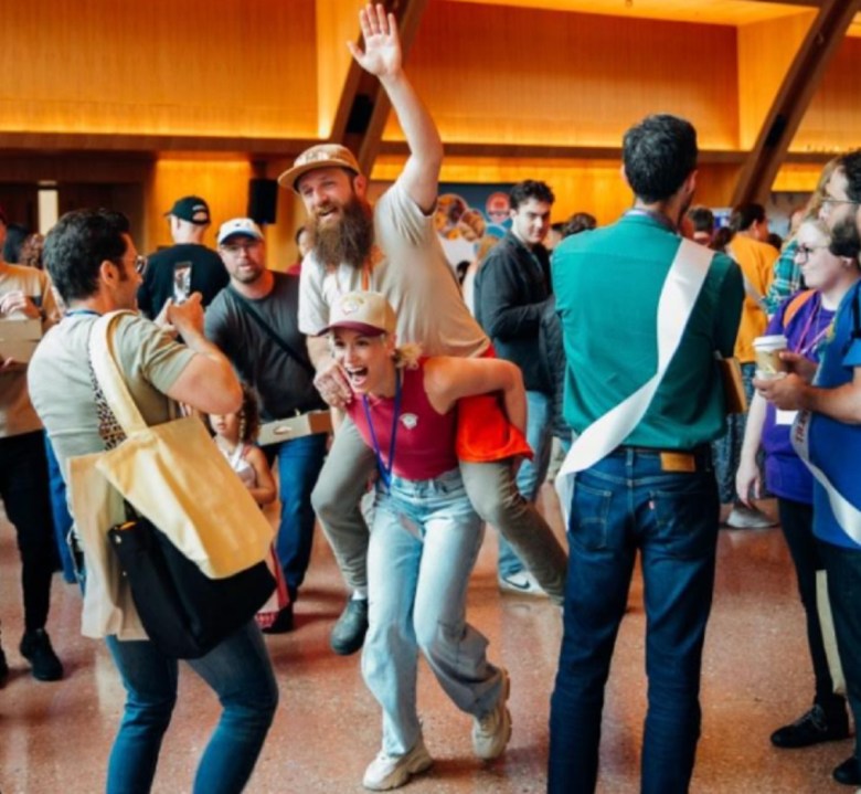 A bearded man is given a piggyback ride by a woman in a crowded room.