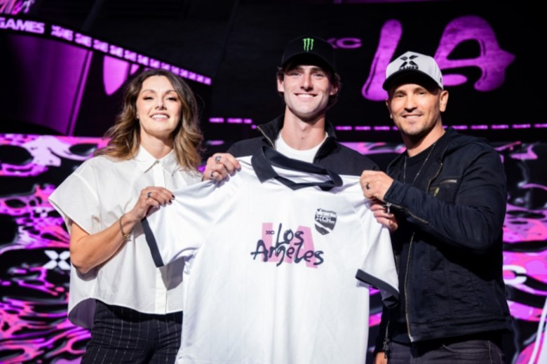 A man joins two others holding his new jersey across his chest on the bright backdrop of a club. It's the X Games League draft party.