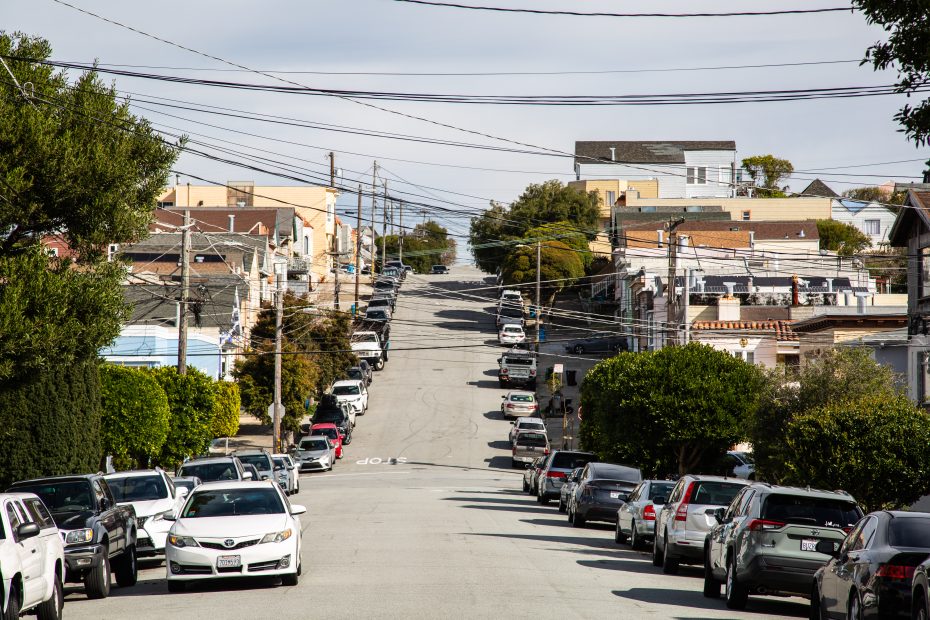 A steep urban street lined with parked cars on both sides, suburban houses, power lines, and trees under a partly cloudy sky.