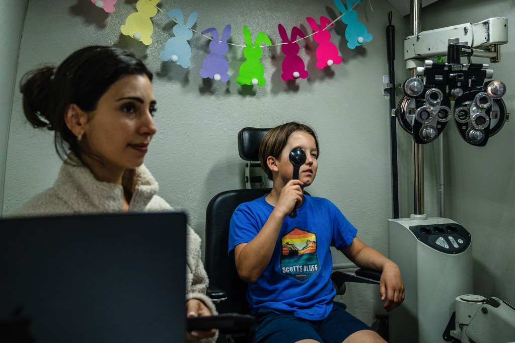 A child places an eye occluder on their right eye and looks forward as a doctor sits next to them during a vision exam.