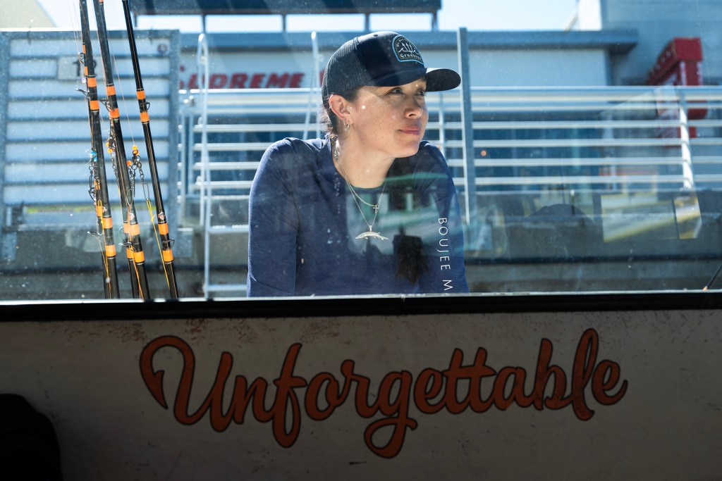 A person wearing a dark long-sleeve shirt and a baseball cap leans against the side of a boat, looking off to the right. Fishing rods stand upright behind them. The scene is viewed through a window, with light reflections and smudges visible on the glass. The word “Unforgettable” is painted in large script along the side of the boat below the window.