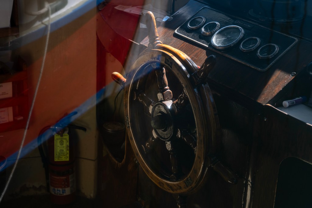 A wooden boat steering wheel sits in the foreground, mounted to a dark console with several round gauges. Warm sunlight filters across the scene, catching the worn varnish of the wheel and creating reflections on nearby surfaces. A fire extinguisher is mounted below, and ropes and storage bins are partially visible in the background, suggesting the interior of a working vessel.