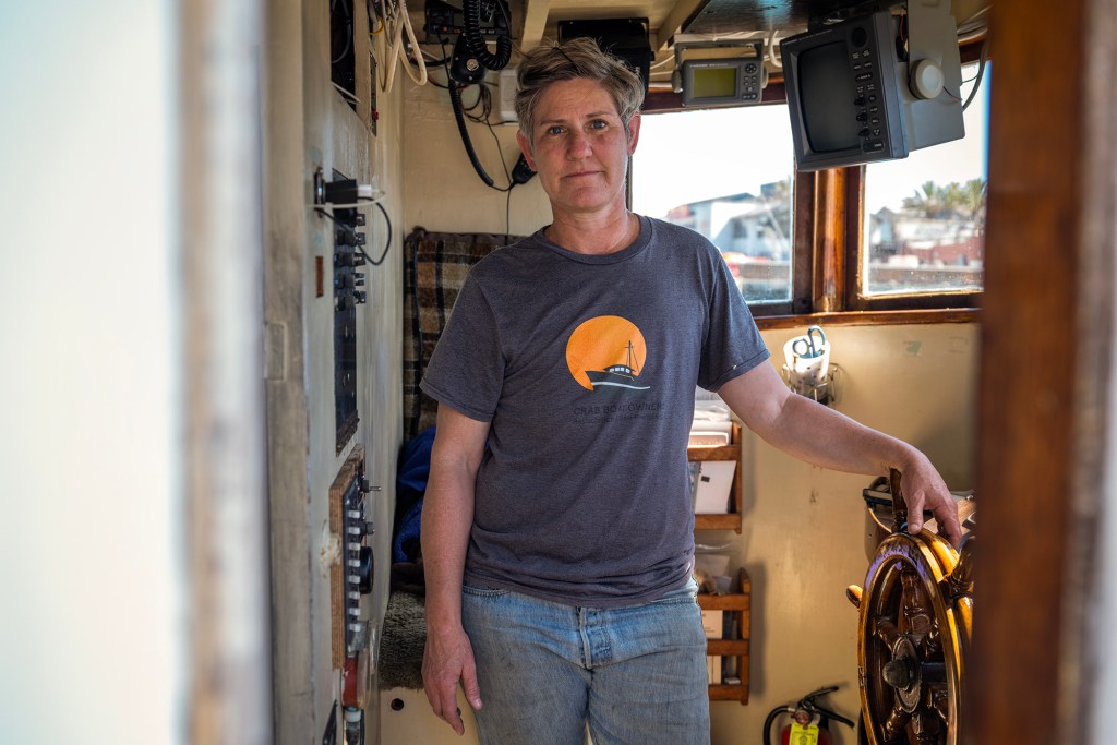 A person stands inside the cabin of a fishing boat, facing the camera with one hand resting on a wooden steering wheel. They wear a gray T-shirt with a boat graphic and light-colored jeans. The compact space is lined with control panels, radios, and navigation equipment, with daylight coming through the windows behind them.