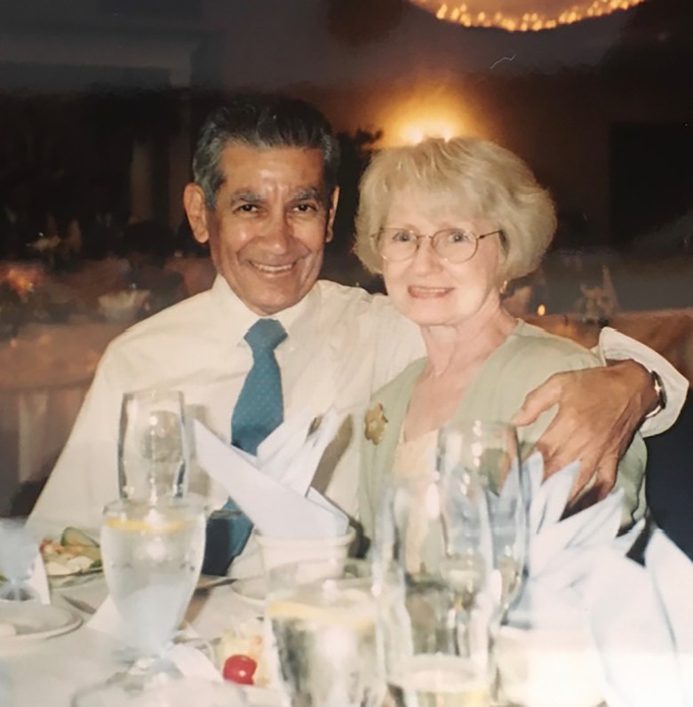 Two people sit closely together at a restaurant table, smiling toward the camera. One has an arm around the other’s shoulder. Glassware, folded napkins and plates are arranged on the table, and warm ambient lighting glows in the background, suggesting an evening dining setting.