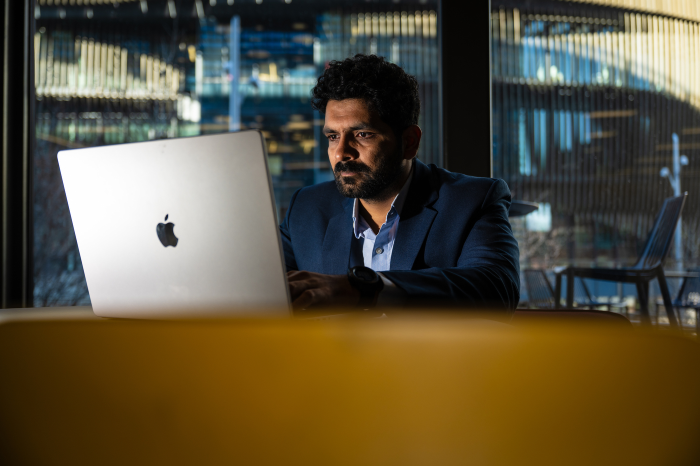 Hitanshu Pandit working at a laptop in front of a row of glass windows. 