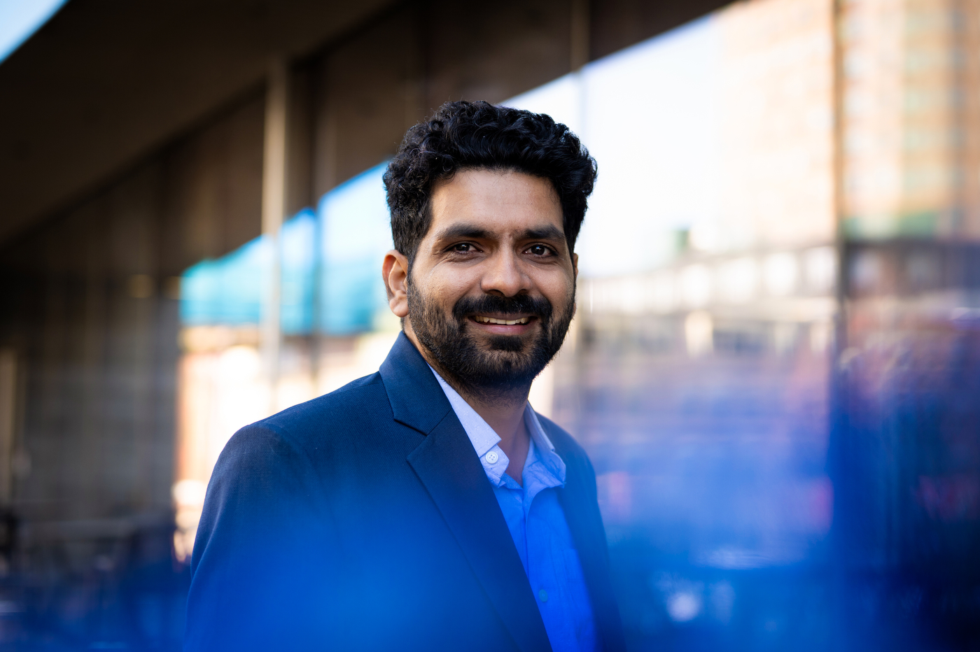 Portrait of Hitanshu Pandit standing outside in front of a wall of windows. 