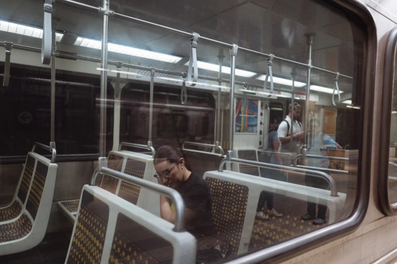 A woman sits alone on a subway train, looking down, with patterned seats and empty handrails around her. The atmosphere is quiet and introspective.
