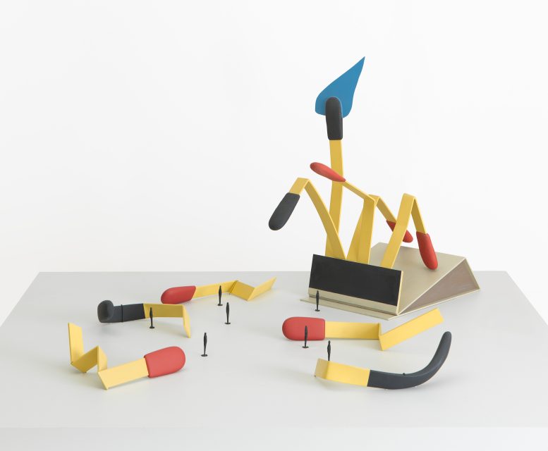 Abstract sculpture with yellow zigzag forms, black and red cylindrical shapes, a blue triangle, and small black pegs on a white surface and background.