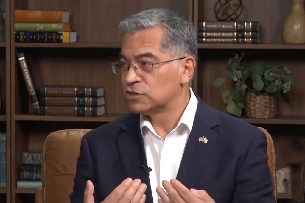 Xavier Becerra speaking in a room with bookshelves and a plant in the background.
