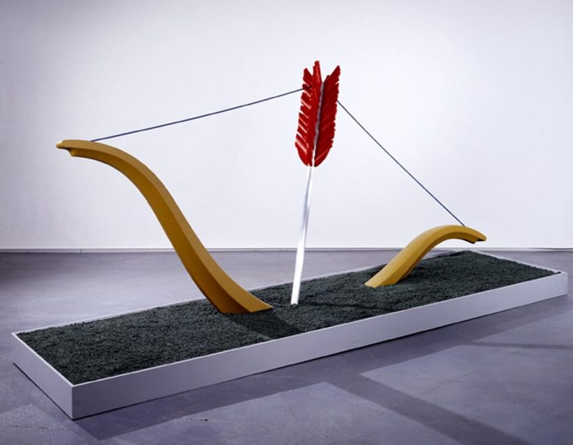 A large sculptural installation of a stylized bow and arrow, with exaggerated curves and a red arrow, displayed on a rectangular platform covered in artificial grass.