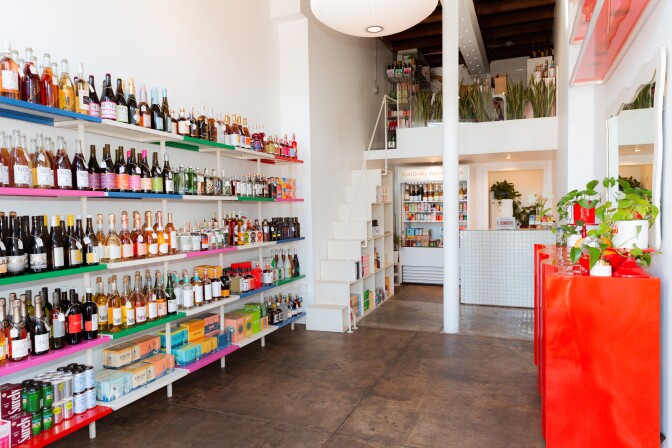An image of a white room thats located inside of a storefront with shelving containing different bottles containing non-alcoholic spirits. There is a polished concrete floor with stairs leading up to a loft area and a red counter on the opposite side of the shelves.  