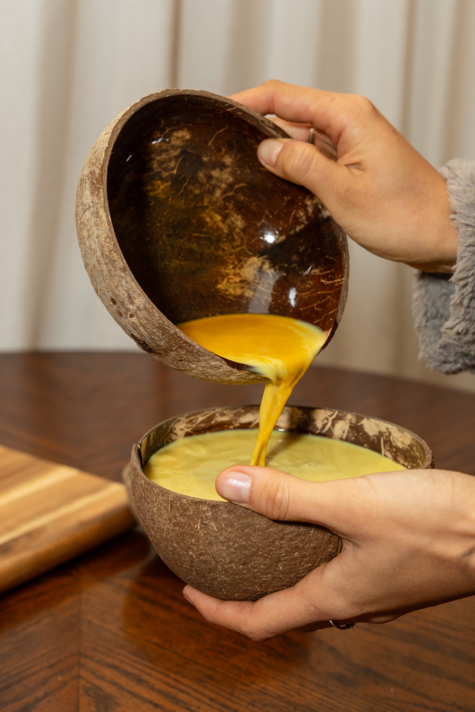 Light brown-skinned hands hold two dark brown bowls resembling coconut shell halves. One hand holds a bowl upright while the other pours a thick golden-yellow liquid into the corresponding bottom bowl