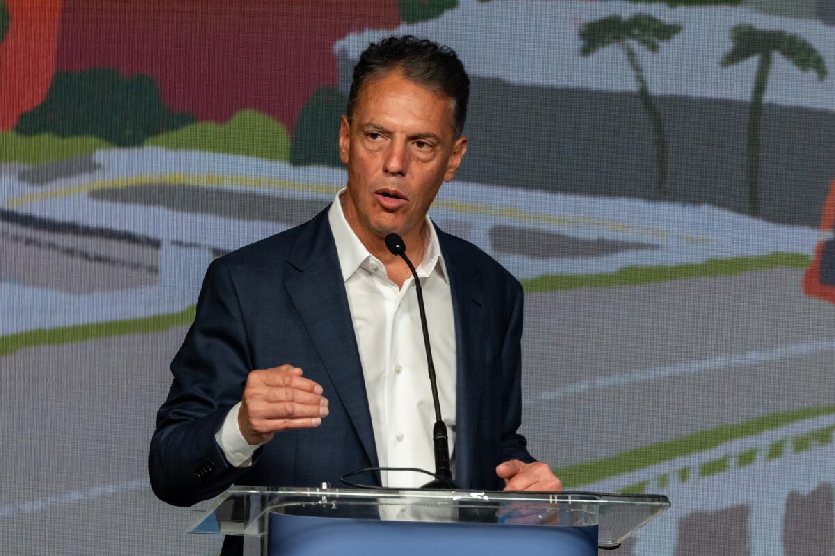 A man with dark hair, in a dark suit and white shirt, gestures while speaking at a lectern