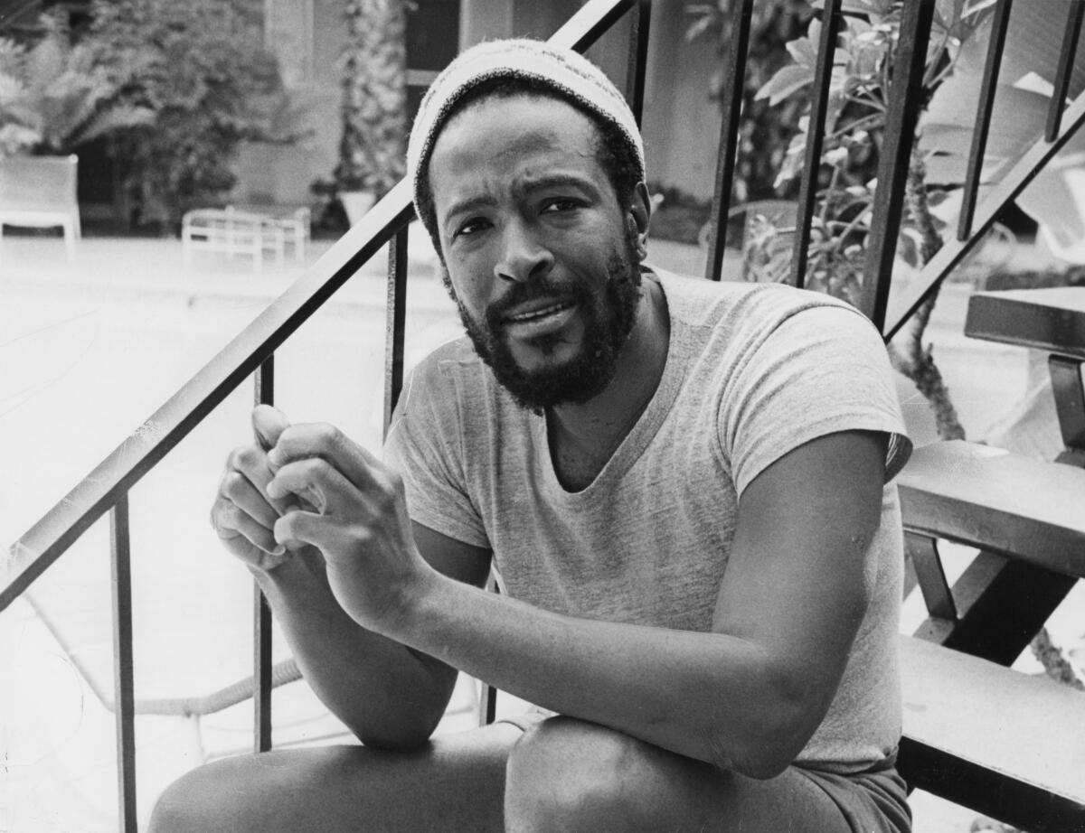 A man with a dark beard, wearing a knit cap, puts his fingers together while seated on a flight of stairs outside